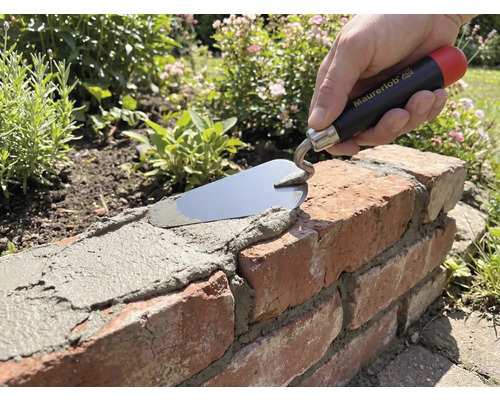 Main avec truelle Maurerlob appliquant du mortier sur un mur en briques dans un jardin.