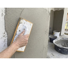 Hand glättet frischen Putz mit einem Reibebrett an einer Wand. Baustelleneimer mit Mörtel im Hintergrund.