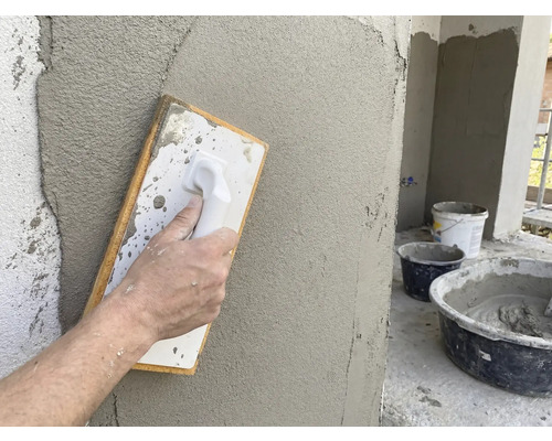 Hand glättet frischen Putz mit einem Reibebrett an einer Wand. Baustelleneimer mit Mörtel im Hintergrund.