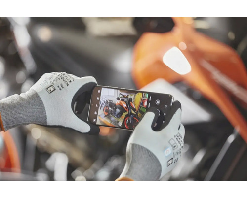 Eine Person trägt Arbeitshandschuhe mit Touch-Funktion und fotografiert ein Motorrad mit einem Smartphone in einer Werkstatt.