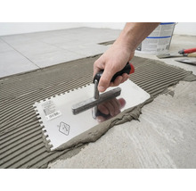 Une main applique de la colle à carrelage sur le sol à l'aide d'une truelle dentelée en formant des rainures.