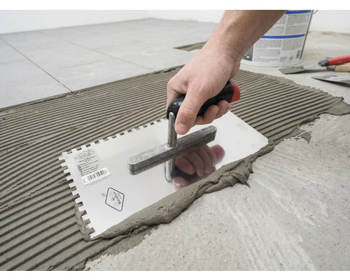 Une main applique de la colle à carrelage sur le sol à l'aide d'une truelle dentelée en formant des rainures.