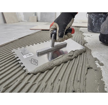 Hand mit Arbeitshandschuh trägt Fliesenkleber mit einer gezahnten Edelstahl-Glättkelle auf den Boden auf.