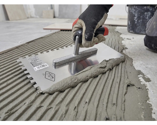 Hand mit Arbeitshandschuh trägt Fliesenkleber mit einer gezahnten Edelstahl-Glättkelle auf den Boden auf.