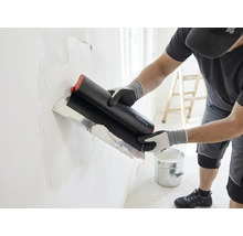 Artisan appliquant de l'enduit sur un mur avec un large couteau à enduire, un seau et une échelle se trouvent dans la pièce lumineuse.