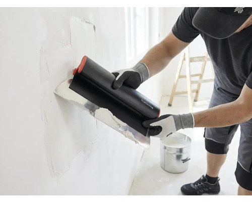 Artisan appliquant de l'enduit sur un mur avec un large couteau à enduire, un seau et une échelle se trouvent dans la pièce lumineuse.