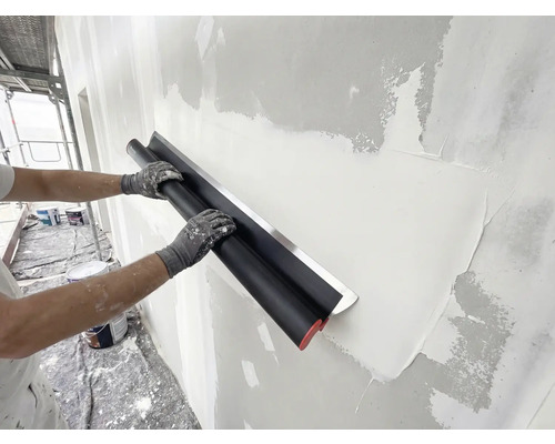 Person glättet Putz an einer Wand mit einem breiten Flächenspachtel aus Edelstahl während Renovierungsarbeiten.