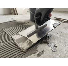 Une personne applique de la colle à carrelage sur un sol en béton avec un peigne à colle en métal.