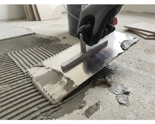 Une personne applique de la colle à carrelage sur un sol en béton avec un peigne à colle en métal.