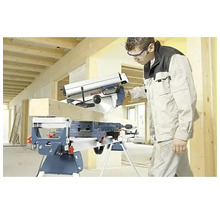 Artisan coupant une poutre en bois avec une scie à onglets Bosch sur un support de travail dans un bâtiment en construction lumineux.