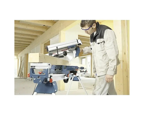 Artisan coupant une poutre en bois avec une scie à onglets Bosch sur un support de travail dans un bâtiment en construction lumineux.