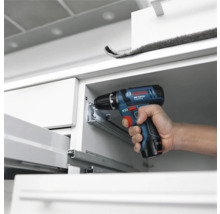 Perceuse-visseuse sans fil Bosch Professional installant des glissières de tiroir en métal dans un meuble blanc.