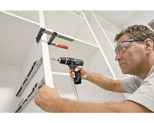 Un homme avec des lunettes de protection monte des glissières de tiroir dans une armoire blanche avec une perceuse-visseuse Bosch sans fil.