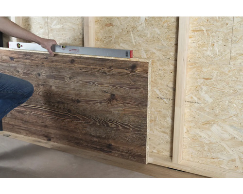 Une personne aligne un panneau mural imitation bois sur un mur en bois avec un niveau à bulle.