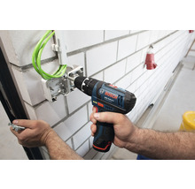 Perceuse-visseuse sans fil Bosch Professional fixant une boîte de dérivation avec câbles verts sur un mur en briques blanches.