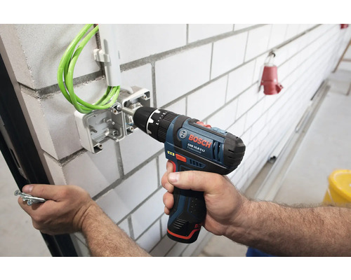 Perceuse-visseuse sans fil Bosch Professional fixant une boîte de dérivation avec câbles verts sur un mur en briques blanches.