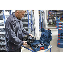 Un homme insère un plateau pour perceuse sans fil dans un coffret bleu Bosch L-BOXX dans un entrepôt organisé avec des étagères.