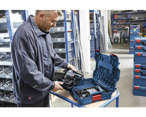 Un homme insère un plateau pour perceuse sans fil dans un coffret bleu Bosch L-BOXX dans un entrepôt organisé avec des étagères.