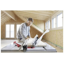 Un homme avec des lunettes de protection coupe une planche de bois avec une scie circulaire sur table dans une pièce lumineuse avec des poutres.