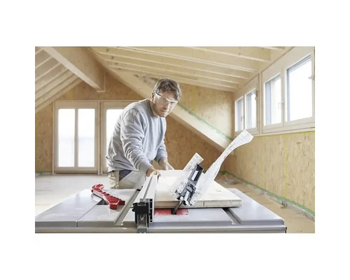 Un homme avec des lunettes de protection coupe une planche de bois avec une scie circulaire sur table dans une pièce lumineuse avec des poutres.