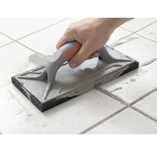 Une main guide une taloche à jointer sur des carreaux de sol blancs pour étaler du mortier de jointoiement gris.