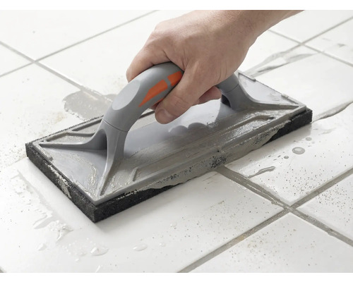 Une main guide une taloche à jointer sur des carreaux de sol blancs pour étaler du mortier de jointoiement gris.