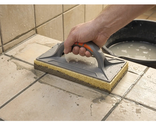 Taloche éponge utilisée pour nettoyer des carreaux fraîchement jointoyés. Une main guide l'outil sur les carreaux mouillés.