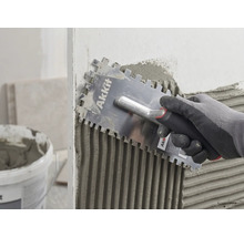 Eine Hand mit Handschuh trägt mit einer Akkit Glättkelle Fliesenkleber gleichmäßig auf eine Wand auf.