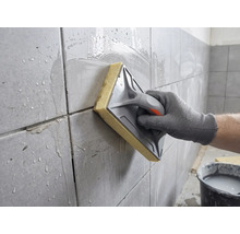 Une main nettoie des carreaux muraux fraîchement jointoyés avec une taloche à éponge et de l'eau.