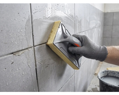 Une main nettoie des carreaux muraux fraîchement jointoyés avec une taloche à éponge et de l'eau.