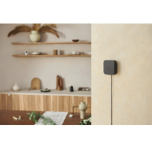 Hub Smart Home noir sur un mur clair dans un intérieur moderne et lumineux avec des étagères en bois et de la décoration.