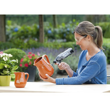 Frau mit Schutzbrille graviert einen Krug aus Terrakotta mit einem Dremel Multifunktionswerkzeug in einem hellen Garten.