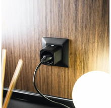 Prise connectée noire insérée dans une prise murale sur un panneau en bois avec un câble d'alimentation branché.