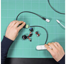 Des mains assemblent différents boutons-poussoirs et composants électroniques sur un tapis de découpe vert pour un projet de bricolage.