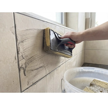 Une personne nettoie des carreaux muraux beiges à l'aide d'une taloche éponge. Un seau blanc rempli d'eau se trouve au premier plan.