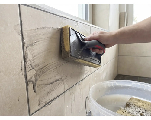 Une personne nettoie des carreaux muraux beiges à l'aide d'une taloche éponge. Un seau blanc rempli d'eau se trouve au premier plan.