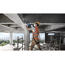 Bauarbeiter mit Schutzausrüstung nutzt Bosch Bohrhammer an einer Betonstruktur auf einer Baustelle mit Gerüst und Stadtansicht.