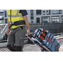 Un ouvrier portant un gilet de sécurité tire un chariot de transport chargé de coffrets à outils Bosch L-BOXX et d'une radio de chantier.