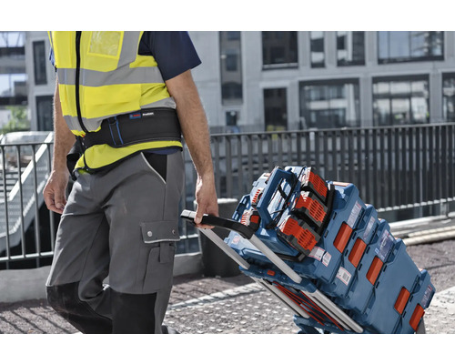 Un ouvrier portant un gilet de sécurité tire un chariot de transport chargé de coffrets à outils Bosch L-BOXX et d'une radio de chantier.