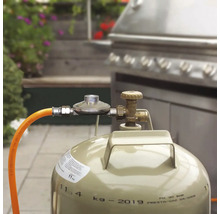 Bouteille de gaz de 11 kilogrammes avec détendeur et tuyau raccordée à un barbecue en acier inoxydable en extérieur.