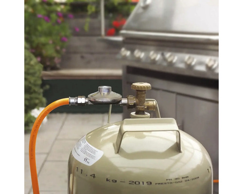 Bouteille de gaz de 11 kilogrammes avec détendeur et tuyau raccordée à un barbecue en acier inoxydable en extérieur.