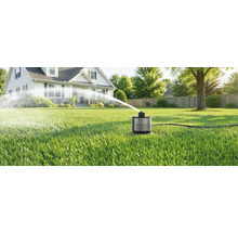 Kompakter smarter Rasensprenger auf grünem Rasen sprüht Wasserbogen vor einem Wohnhaus im hellen Garten.