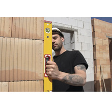 Un ouvrier du bâtiment portant un casque de protection blanc utilise un niveau à bulle jaune pour vérifier l'alignement d'un mur en briques.