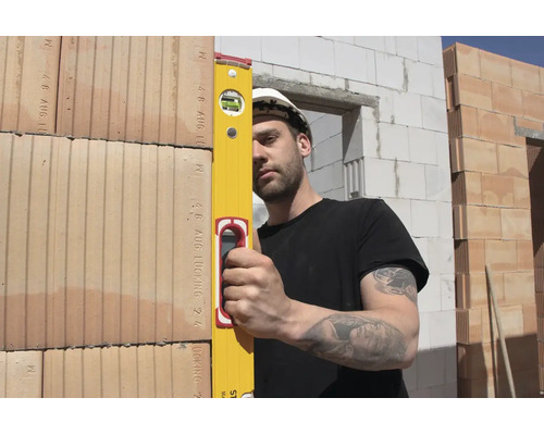 Un ouvrier du bâtiment portant un casque de protection blanc utilise un niveau à bulle jaune pour vérifier l'alignement d'un mur en briques.