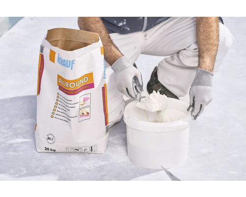 Un artisan prélève de l'enduit dans un seau à côté d'un sac de 20 kilogrammes d'enduit universel Knauf Allround 2 en 1.