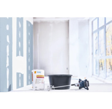 Un sac de 20 kilogrammes d'enduit Knauf Allround à côté d'un seau à mélanger noir et d'un malaxeur électrique sur un chantier lumineux.