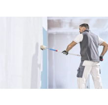 Artisan en vêtements de travail peignant un mur avec un rouleau à peinture et une perche télescopique dans un intérieur lumineux.