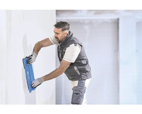 Un artisan lisse un mur blanc avec un large couteau à enduire bleu dans une pièce lumineuse.