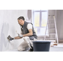 Un artisan en vêtements de travail applique de l'enduit sur un mur avec une lisseuse et un couteau à enduire.