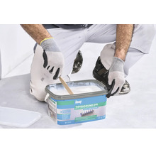 Une personne s'agenouille et mélange le gel de fond blanc Knauf dans un seau bleu avec un bâton en bois.